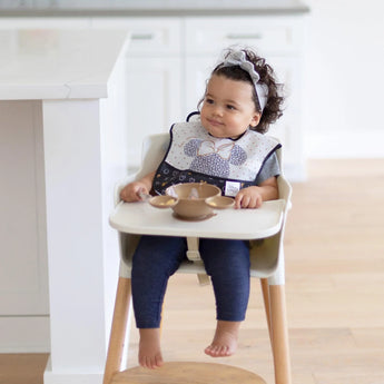 A toddler wearing a bib and headband with Disney mouse ears sits ready in their highchair, using the Bumkins Minnie Mouse Gold Feeding Set.