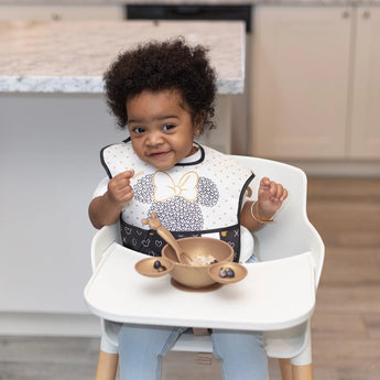 A curly-haired toddler sits in a highchair, wearing a bib and using Bumkins Silicone First Feeding Set: Minnie Mouse Gold, smiling while eating.