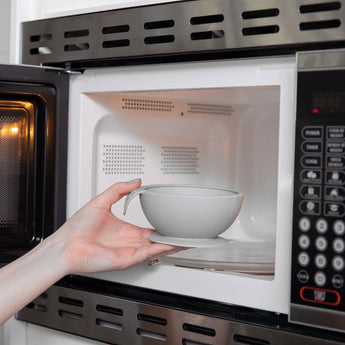 A persons hand places a gray bowl from the Bumkins Silicone First Feeding Set into an open microwave. The microwaves right-side digital control panel carefully heats your food-safe silicone items.