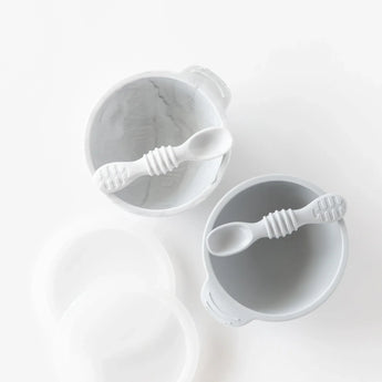 Two gray Bumkins Silicone First Feeding Set bowls with handles, matching baby spoons inside, and removable lids on a light surface.