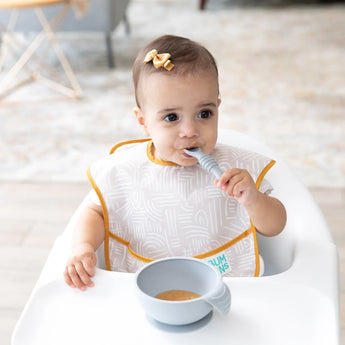 A baby with a bow sits in a high chair, holding their Bumkins silicone spoon from the gray feeding set, ready for self-feeding.