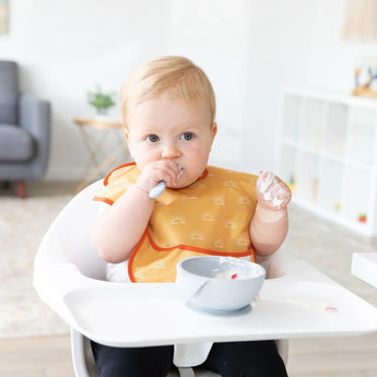 Baby with an orange bib self-feeds using Bumkins Silicone First Feeding Set: Gray. In the background, a chair and shelves are visible.