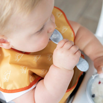 Baby self-feeds using a Bumkins Silicone First Feeding Set in Gray, wearing a patterned bib.