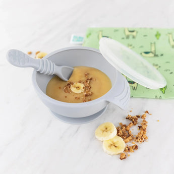 A bowl of banana puree with granola, slices, and a Bumkins Silicone First Feeding Set baby spoon sits on a marble surface.