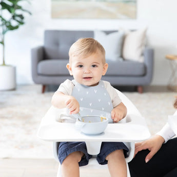 A toddler in a high chair uses the Bumkins Silicone First Feeding Set: Gray while holding a bowl and baby spoon with an adult hand nearby.