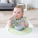 Baby wearing a colorful bib with the Bumkins Silicone First Feeding Set in Gray, holding a spoon in a high chair. Cozy living room backdrop.