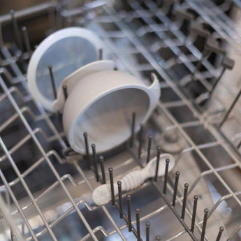 A Bumkins First Feeding Set, a baby spoon, and toothbrush are on the top rack of an open dishwasher.