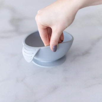 A hand reaches into a Bumkins Silicone First Feeding Set: Gray bowl with a baby spoon on the marble surface.