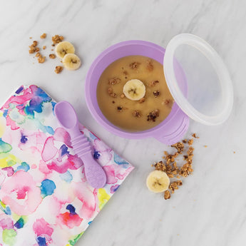 Lavender Bumkins Silicone First Feeding Set with banana oatmeal, granola, and vibrant floral napkin on a marble surface.