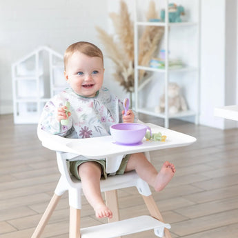 A baby in a high chair holds Bumkins’ Silicone First Feeding Set: Lavender, with a bowl on the tray, smiling in a bright room.