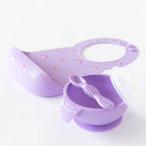 The Bumkins Silicone First Feeding Set: Lavender includes a rainbow-patterned bib, a bowl with lid, and a silicone spoon.