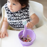 A child in a high chair uses Bumkins Lollipop silicone spoon, holds a purple bowl of oatmeal, and wears a black and white bib.