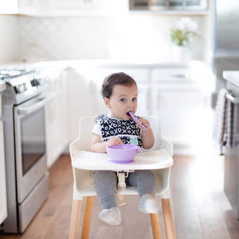 A baby in a high chair uses Bumkins Silicone Dipping Spoons while wearing a patterned bib in a bright, modern kitchen.