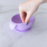 A hand with painted nails holds Bumkins Silicone First Feeding Set: Lavender, a food-grade bowl and spoon, on a light surface.