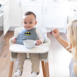 A baby in a high chair, wearing a Batman bib, eagerly reaches for the Bumkins Silicone Dipping Spoon in Taffy.