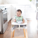 Baby in a high chair, smiling with Bumkins Silicone Dipping Spoons (Taffy) and bowl, wearing a green bib in the bright kitchen.