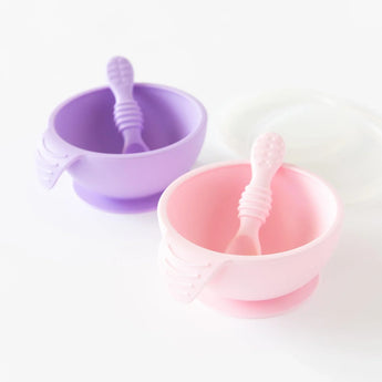 Two Bumkins Silicone First Feeding Sets, in pink and purple, each with a suction base and spoon, displayed on a white surface.