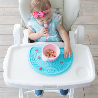 A toddler with a pink bow enjoys cereal and strawberries from the Bumkins Silicone First Feeding Set: Pink, on a blue silicone mat.