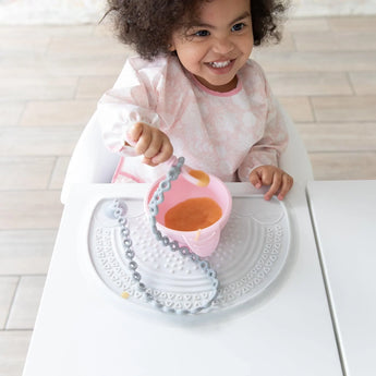 A curly-haired child beams while sitting at a table, holding a utensil and the Bumkins Silicone First Feeding Set: Pink—a bowl filled with food. The table features a food-safe silicone mat and teething toy, promoting self-feeding adventures.