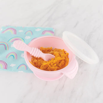 A Silicone First Feeding Set: Pink by Bumkins with orange mash and spoon is on marble, lid ajar next to a rainbow-patterned cloth.