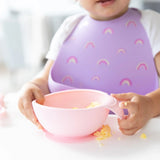 A baby self-feeds from a Bumkins Silicone First Feeding Set: Pink with a purple bib and rainbows at a white table.