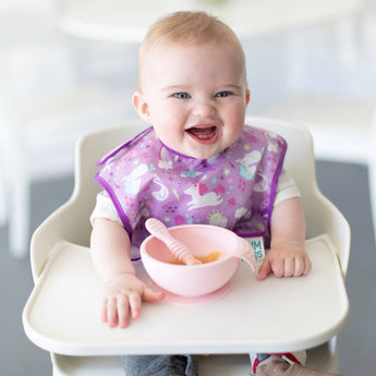 In a softly blurred background, a smiling baby sits in a high chair wearing a purple unicorn bib. The baby practices self-feeding with Bumkins Silicone First Feeding Set: Pink, holding the food-safe silicone bowl and spoon, adding to the charming scene.