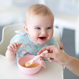 A smiling baby eagerly sits in a high chair, exploring self-feeding with Bumkins Silicone Dipping Spoons from a pink bowl.