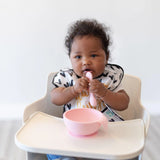 A baby in a high chair with Bumkins Silicone First Feeding Set: Pink, featuring a pink spoon, bib with black hearts, and suction bowl.