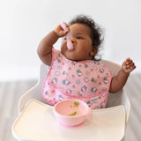 Baby self-feeding in a high chair with Bumkins Silicone Dipping Spoons in Lollipop color, matching a pink bowl and bib.