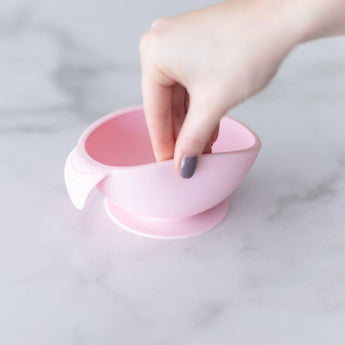 A hand reaches into Bumkins Silicone First Feeding Set: Pink, featuring a small pink bowl with suction base on marble.