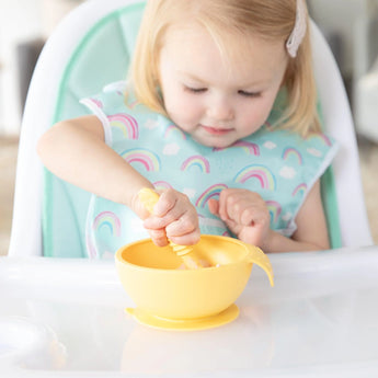 A child in a high chair uses Bumkins silicone dipping spoons from the Tutti-Frutti pack to stir food in a yellow bowl.