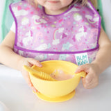 A child uses Bumkins Tutti-Frutti Silicone Dipping Spoon with a unicorn bib, mastering self-feeding at the table.