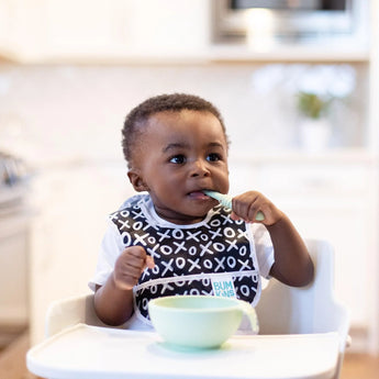 A toddler in a high chair wears a bib, holds a Bumkins Silicone Dipping Spoon with a green bowl in front of them.