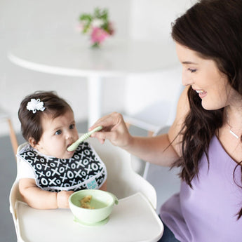 A woman smiles as the baby, in a patterned bib and bow, self-feeds with Bumkins Silicone Dipping Spoons from the Taffy 3 Pack.