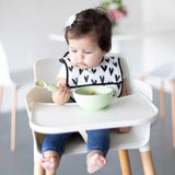 A baby in a high chair uses Bumkins Silicone Dipping Spoons with a heart-patterned bib and a bowl in front.