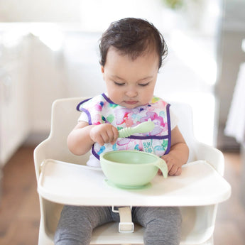 A baby sits in a high chair, wearing a bib, holding Bumkins Silicone Dipping Spoons and curiously eyeing the bowl on the tray.