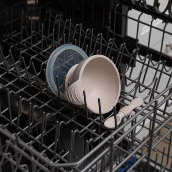 The Bumkins Silicone First Feeding Set: Sand, with a bowl, lid, and spoon, fits perfectly in an open dishwasher rack.