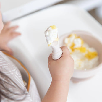 A child practices self-feeding confidently with a Bumkins Silicone First Feeding Set in Sand, holding a spoon near a bowl on the table.