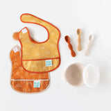 The Bumkins Silicone First Feeding Set in Sand includes two orange bibs, three textured spoons, and a white bowl for safe self-feeding.