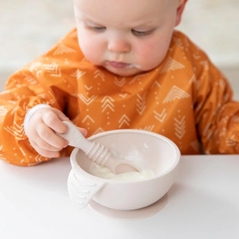 Baby in a Bumkins Silicone First Feeding Set: Sand, joyfully self-feeding yogurt with an orange bib and a smeared face.