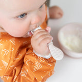 In a Sand silicone feeding set by Bumkins, a baby in an orange bib joyfully self-feeds yogurt with a white spoon.