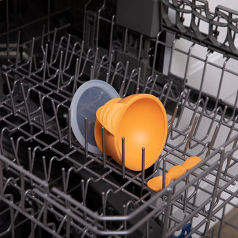 The Bumkins Silicone First Feeding Set in Tangerine is secure on the top rack of a gray dishwasher, cup and lid upside down.