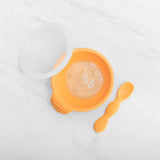 Bumkins Silicone First Feeding Set in Tangerine, with suction base, spoon, and lid; filled with puree on a white surface.