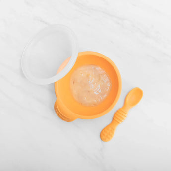 Bumkins Silicone First Feeding Set in Tangerine, with suction base, spoon, and lid; filled with puree on a white surface.