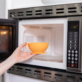 A hand places the Bumkins Silicone First Feeding Set: Tangerine on a rotating glass plate inside an open microwave.