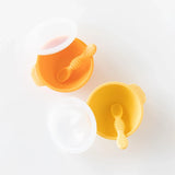 Two Silicone First Feeding Sets by Bumkins, in Tangerine and a matching Yellow, include bowls with lids, spoons & suction bases.