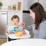 A smiling woman uses the Bumkins Silicone First Feeding Set: Tangerine, feeding a baby in a high chair with a dinosaur bib.