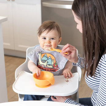 A parent uses a Bumkins Silicone Dipping Spoon from the Tutti-Frutti pack to feed a happy baby in a colorful bib with an orange bowl.