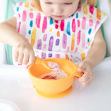 Child self-feeding using Bumkins Silicone First Feeding Set in Tangerine, wearing a colorful bib with abstract patterns.