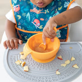 A child in a bib uses Bumkins Silicone First Feeding Set: Tangerine to self-feed with an orange spoon and bowl on the highchair tray.